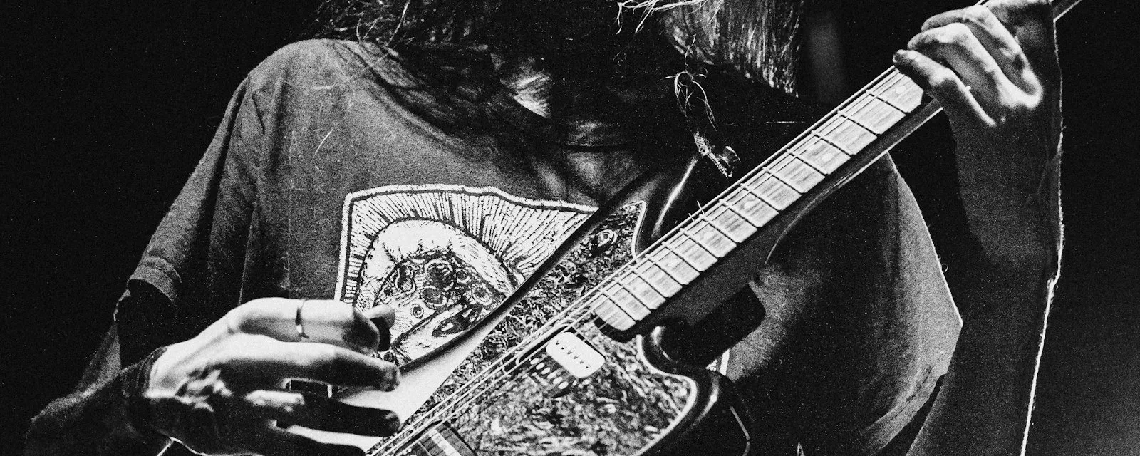 A guitarist playing an electric guitar in high-contrast monochrome, the instrument bright against a near-black background